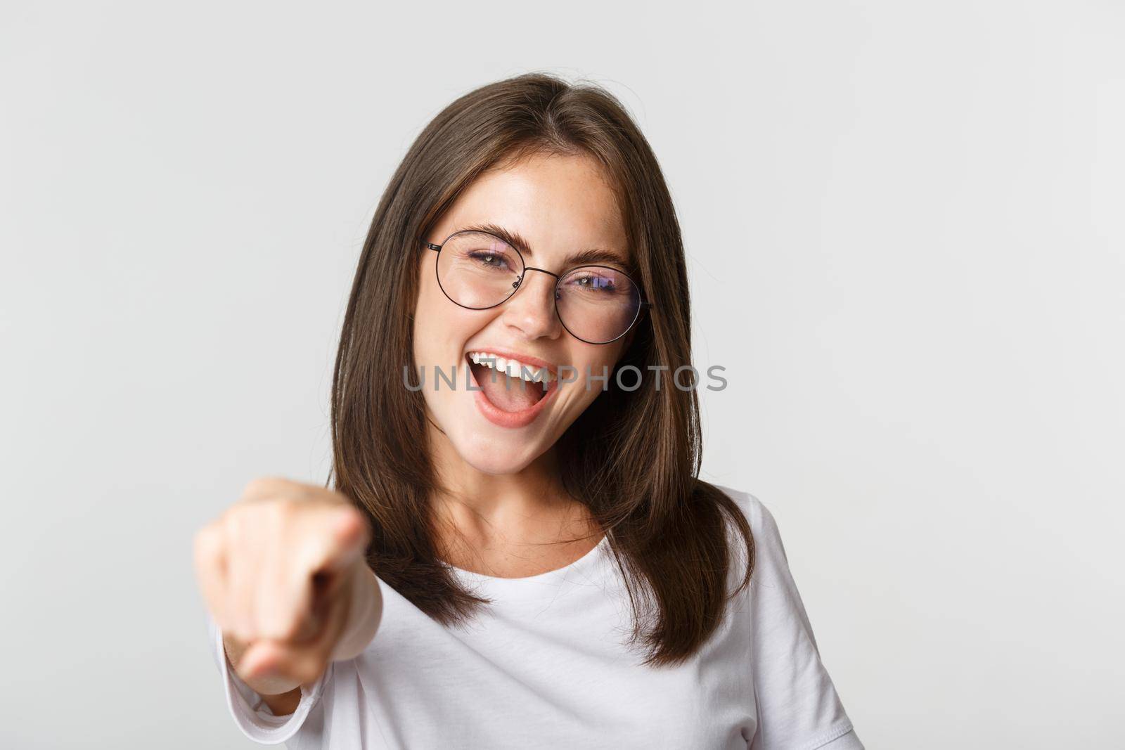 Close-up of cheerful beautiful girl in glasses smiling, poiting finger at camera by Benzoix