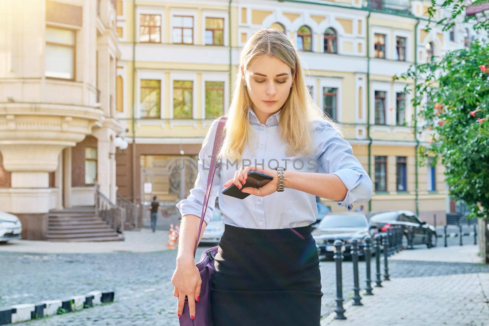 Young business woman in city looking at her wristwatch. Confident serious female with smartphone in her hands, with laptop bag, sunset in the city, urban style background