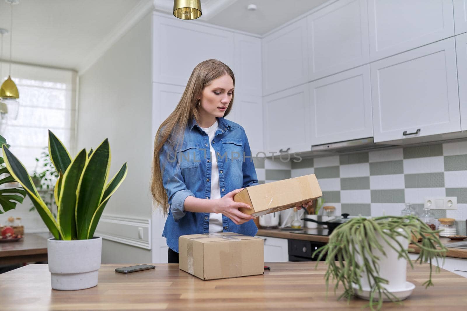 Young happy woman unpacking cardboard boxes, unboxing expected postal parcel with online shopping, at home in kitchen