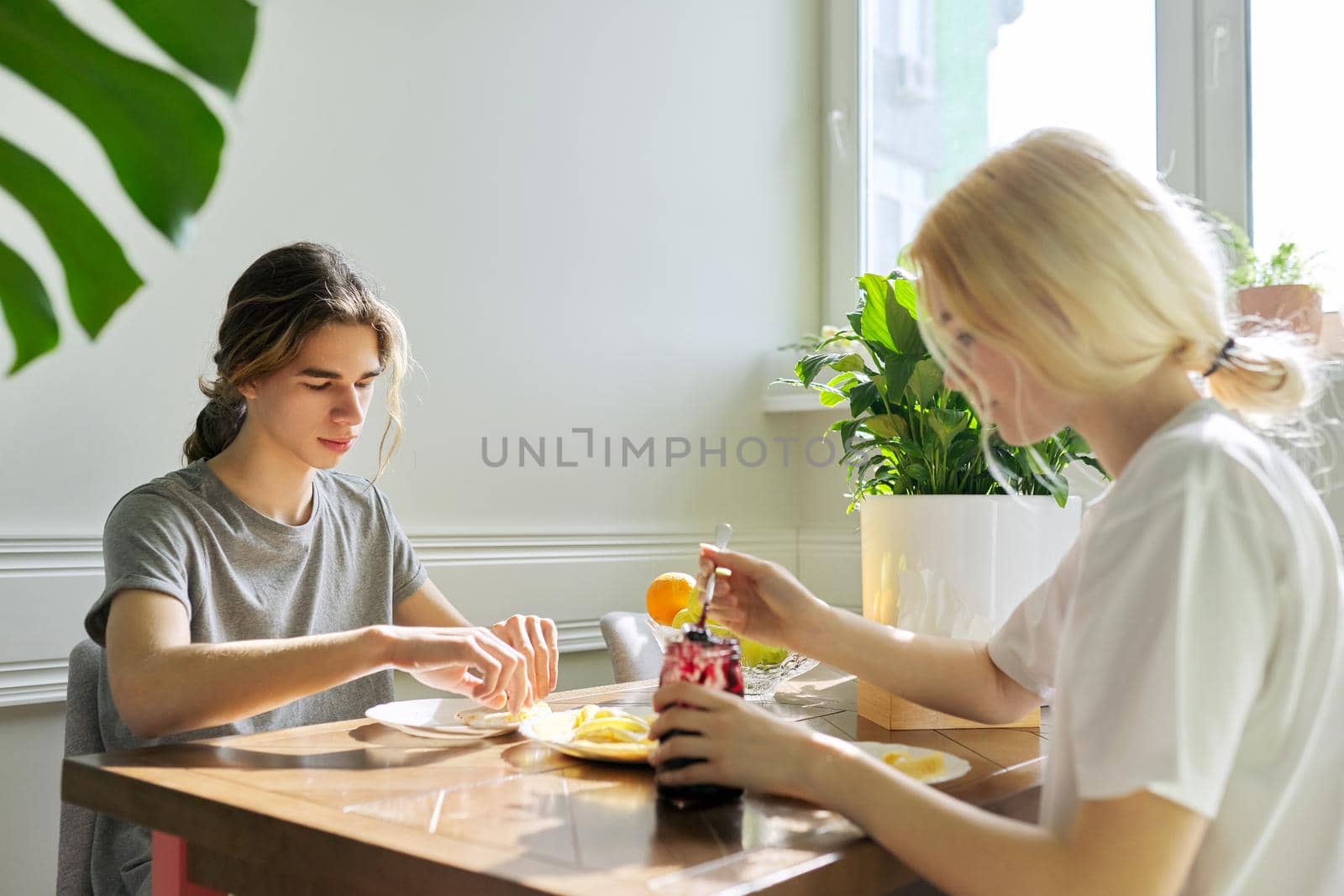 Teenagers guy and girl eating pancakes with jam, sitting at table at home. Homemade food, communication, teens, lifestyle concept