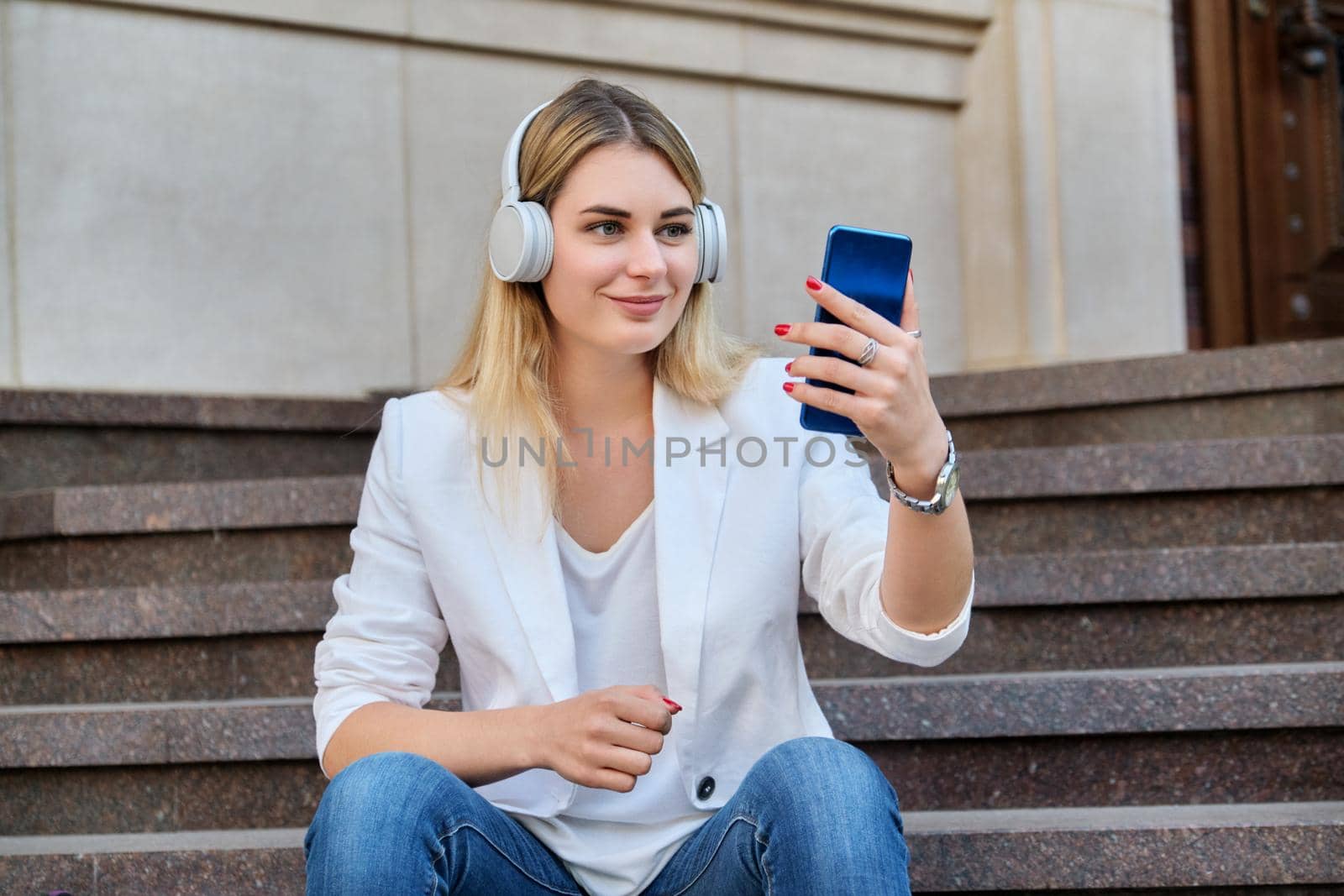 Young beautiful woman in headphones with smartphone resting sitting on steps in city, listening music watching video. Happiness, relaxation, lifestyle, youth and technology