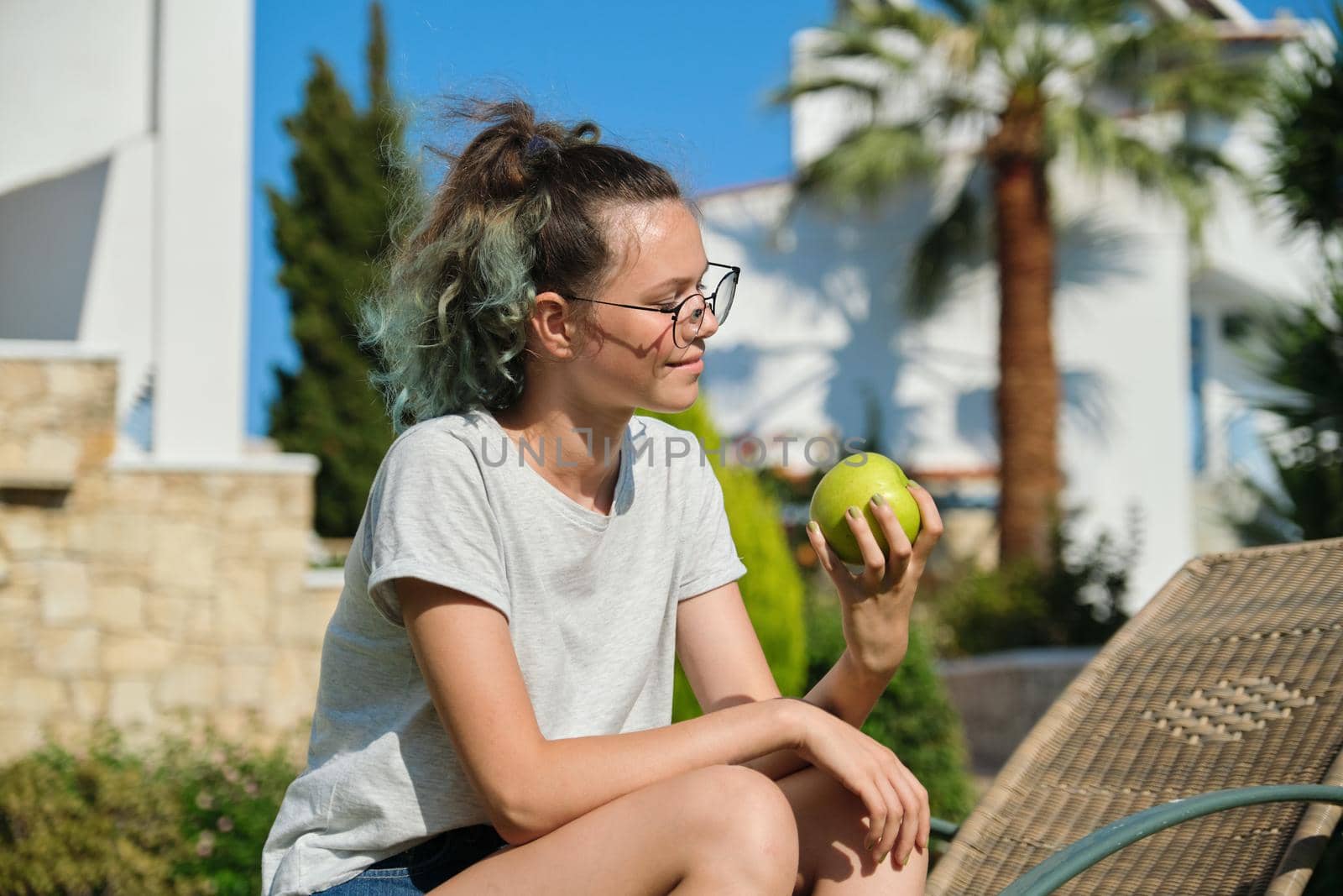 Girl teenager with green apple, resting sitting on sun lounger in garden, female holding tasty healthy apple in hand, sunny summer day grass background