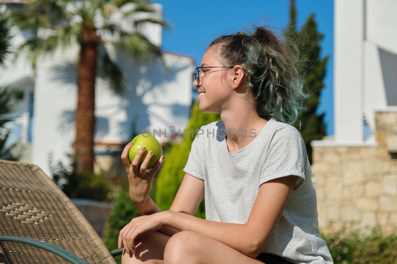 Girl teenager with green apple, resting sitting on sun lounger in garden, female holding tasty healthy apple in hand, sunny summer day grass background