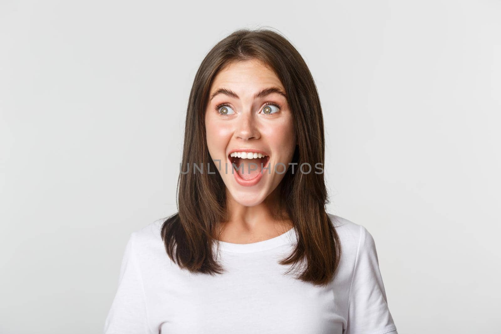 Portrait of joyful pretty brunette girl looking left amused, smiling excited and surprised over white background.