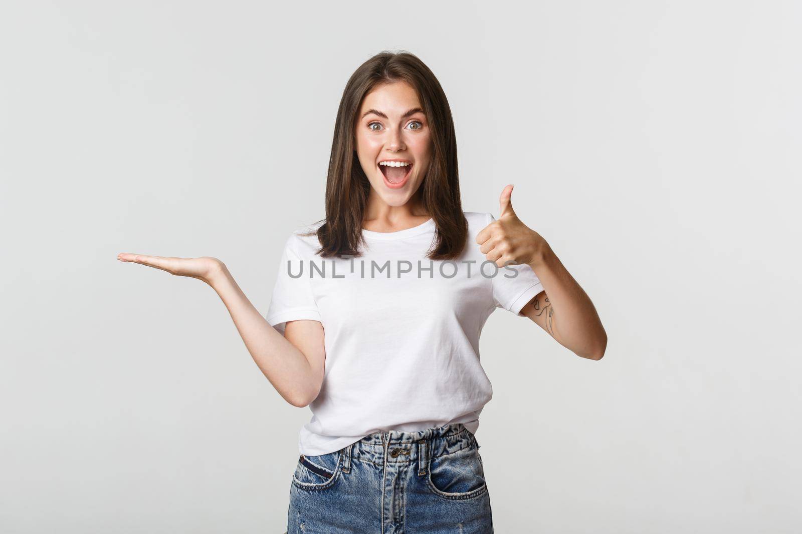 Satisfied attractive girl holding something on hand and showing thumbs-up in approval.