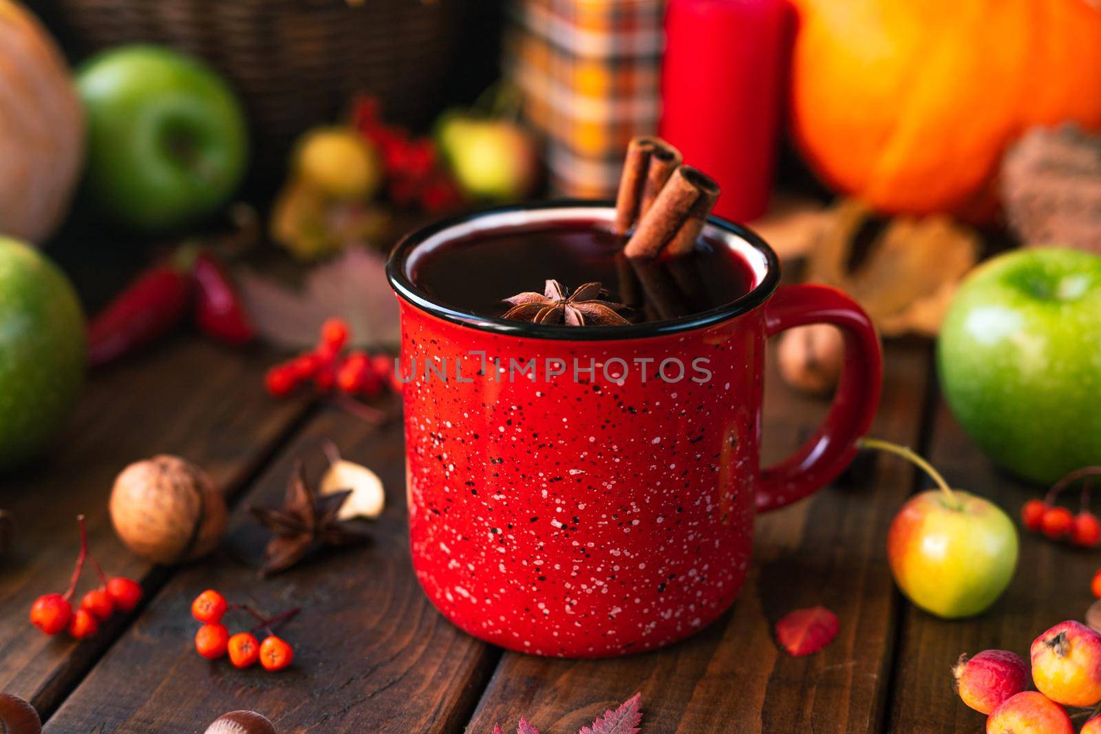 red mug with mulled wine. Cinnamon sticks stick out of the cup and a star of star anise floats. Fruits and spices are all around on a wooden table by Adriablack