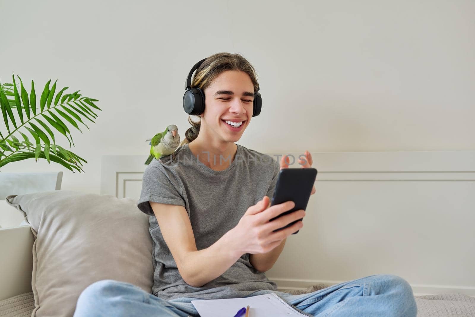 Emotional talking male teenager in headphones looking at smartphone screen using video call. Bird pet green quaker parrot on the owners shoulder, home room sofa bed background