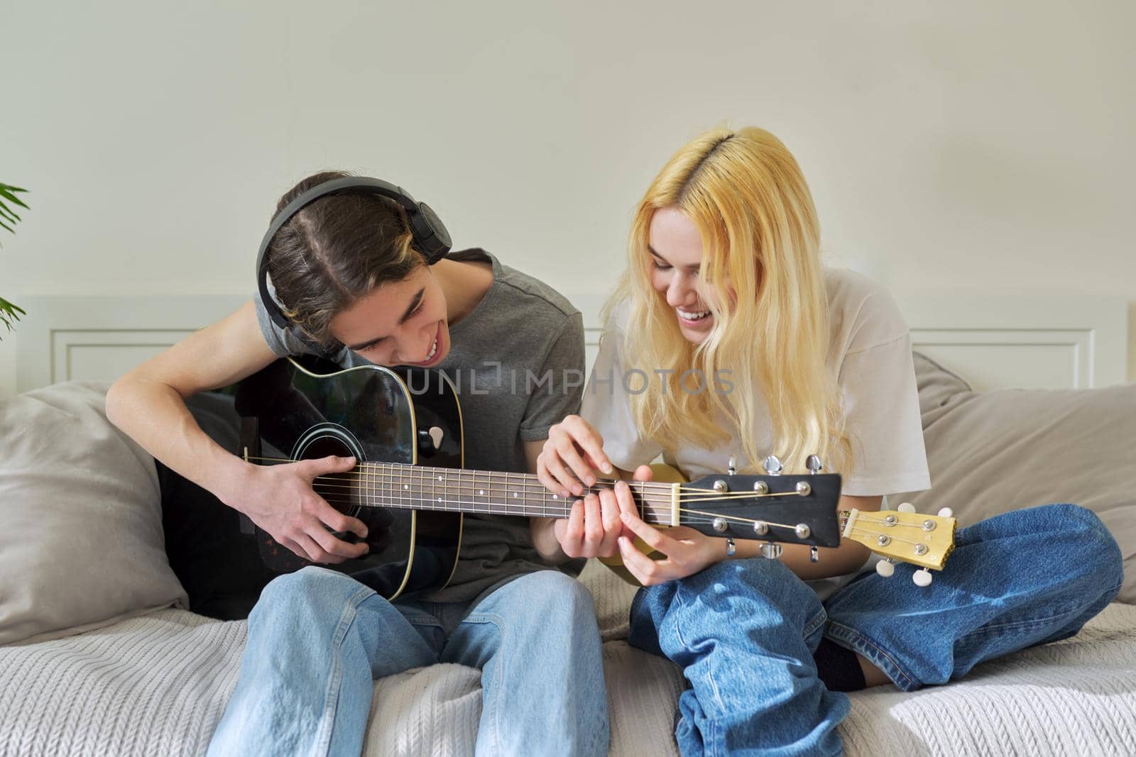 Creative couple of teenagers friends with musical instruments sitting at home on bed. Acoustic guitar and ukulele in hands of guy and girl. Hobby, music, friendship, creativity, youth concept