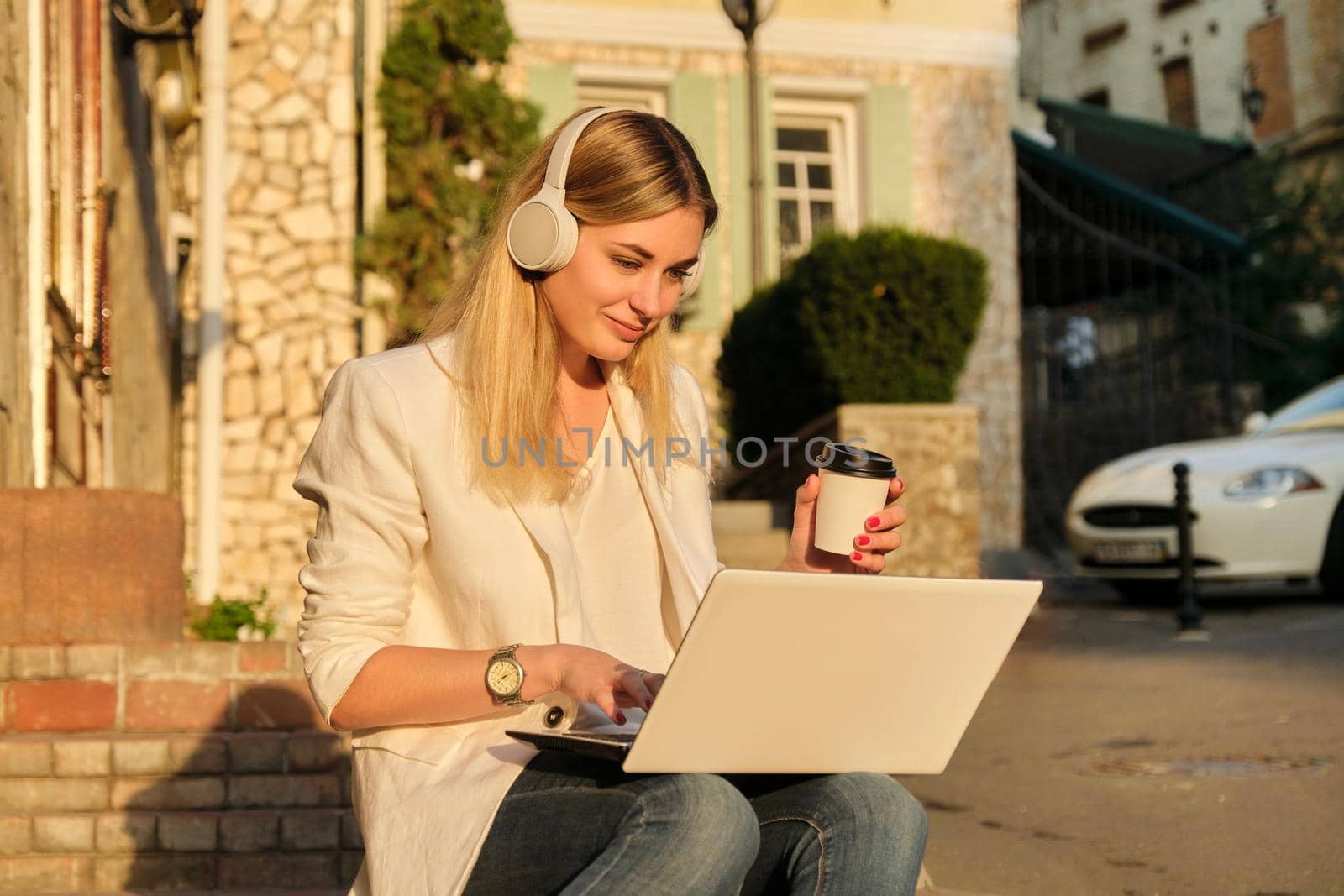 Young beautiful female in headphones with laptop drinking coffee in paper cup. Outdoor portrait of smiling woman sitting on steps in city