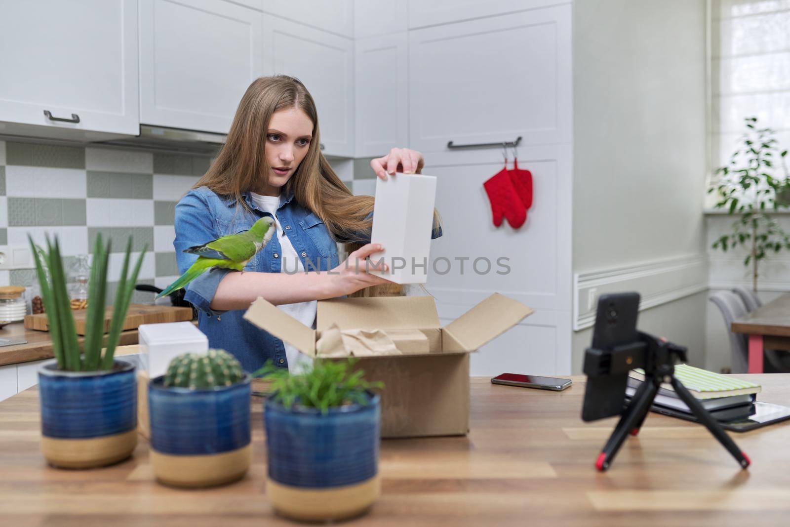 Woman recording video on smartphone moment of unpacking cardboard box. Showing purchases ordered on Internet, product quality reviews with followers, blog content, feedback. Pet parrot helping unpack