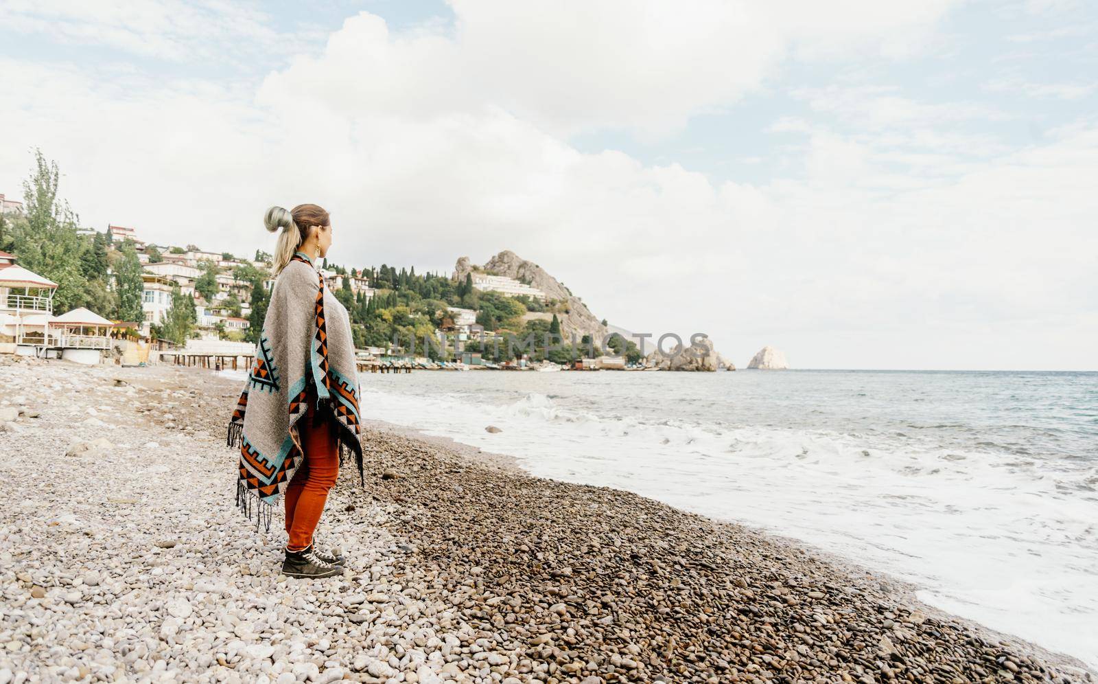 Fashionable boho style young woman standing on pebble beach and looking at sea.