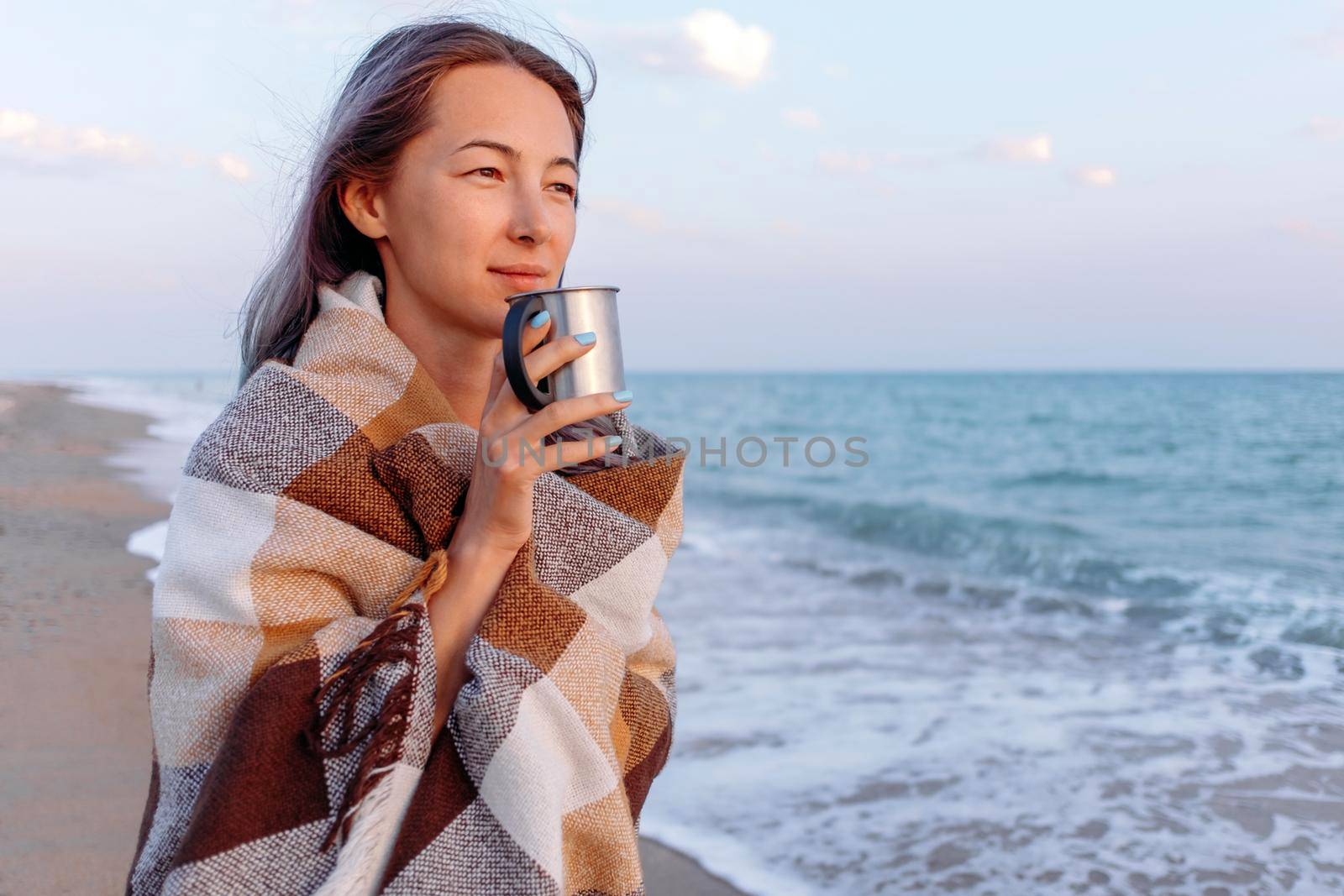 Beach vacation. Young woman wrapped in plaid drinking tea from cup on coast near the sea.