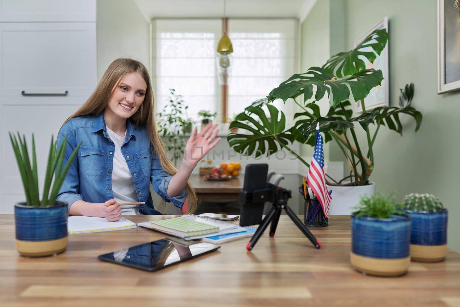 Young woman university student studying at home online remotely, e-learning. Talking female looking at smartphone screen, USA flag background, distance concept