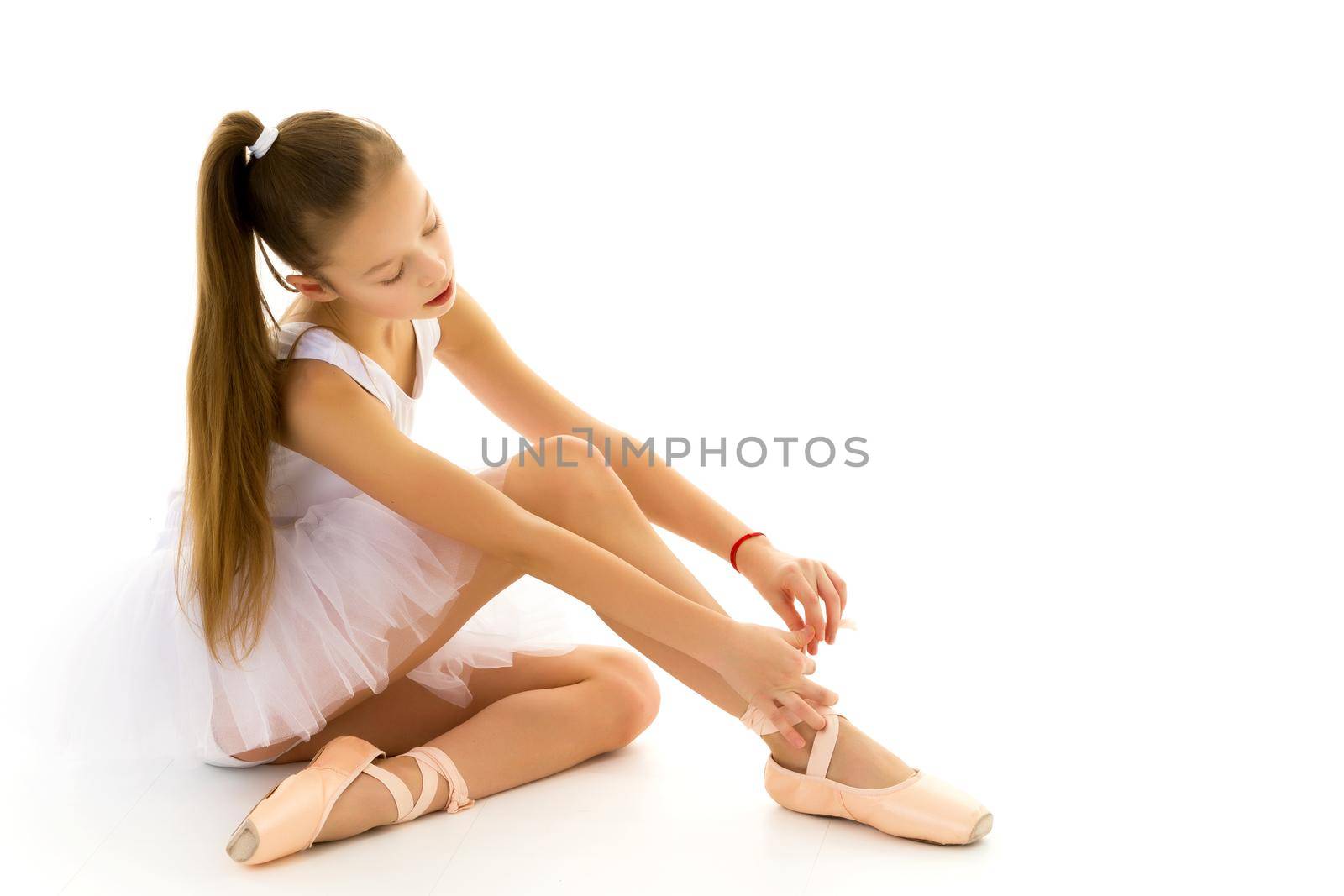 Adorable little girl putting on her ballet dancing shoes, sitting on the floor at a dance studio. Little ballerina is preparing for classes in a ballet school, copy space. Achievement, concept of dedication