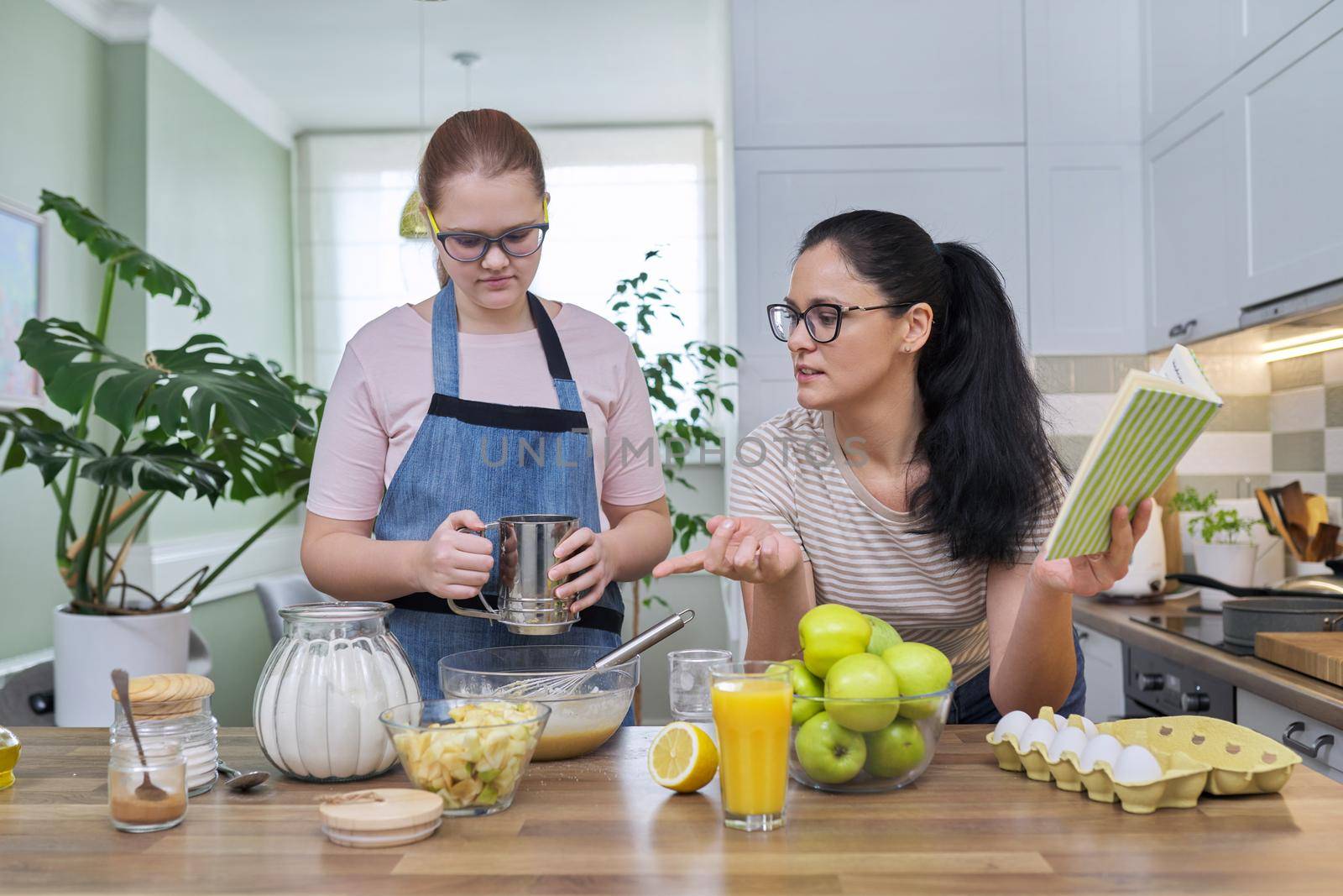 Mom and preteen daughter cooking together at home in kitchen apple pie, mother with book of recipes reading sequence of baking. Family, lifestyle, parent child relationship, homemade food concept