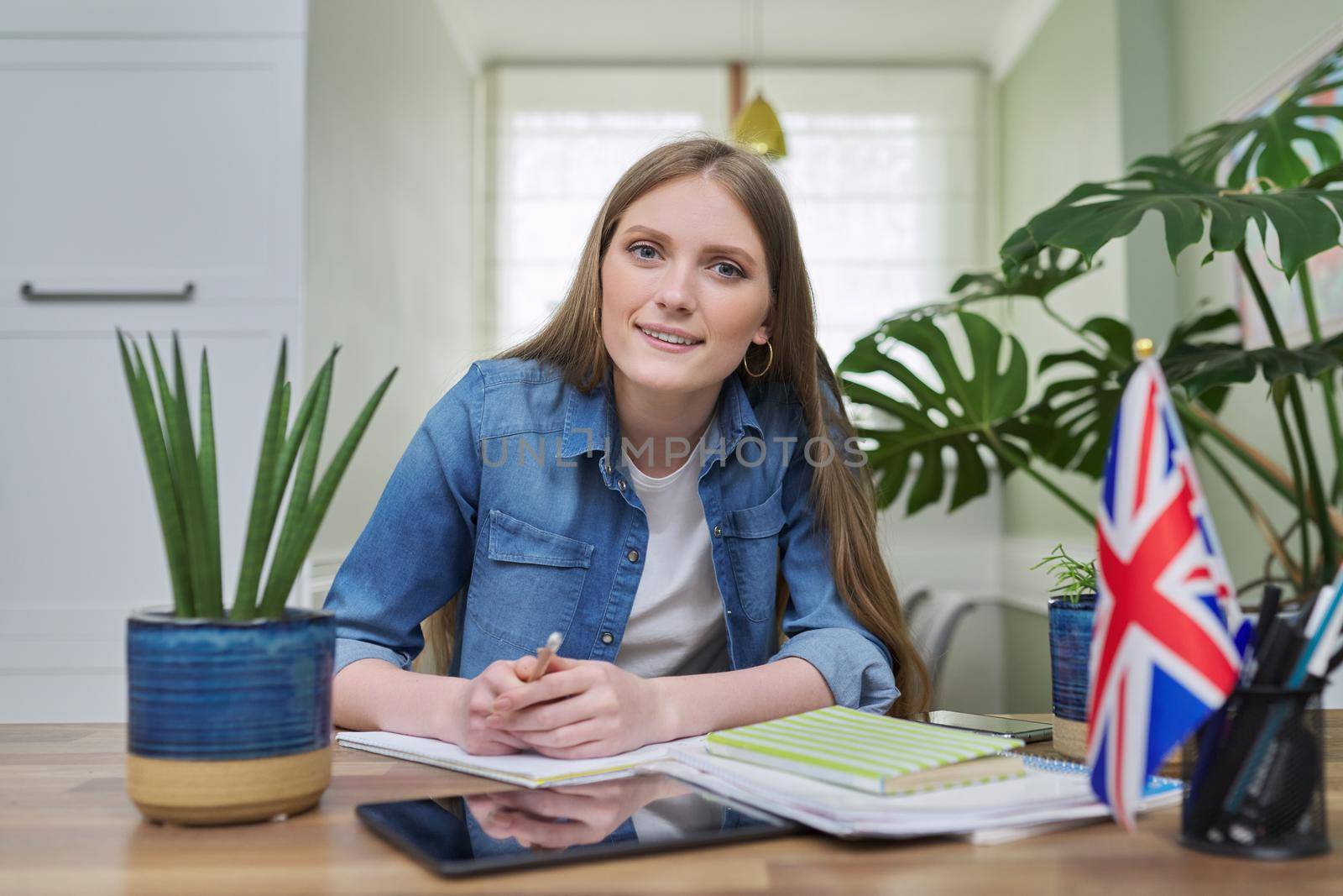 UK learning English online. Female student remotely looking at webcam taking private lessons sitting at home in kitchen, England flag background