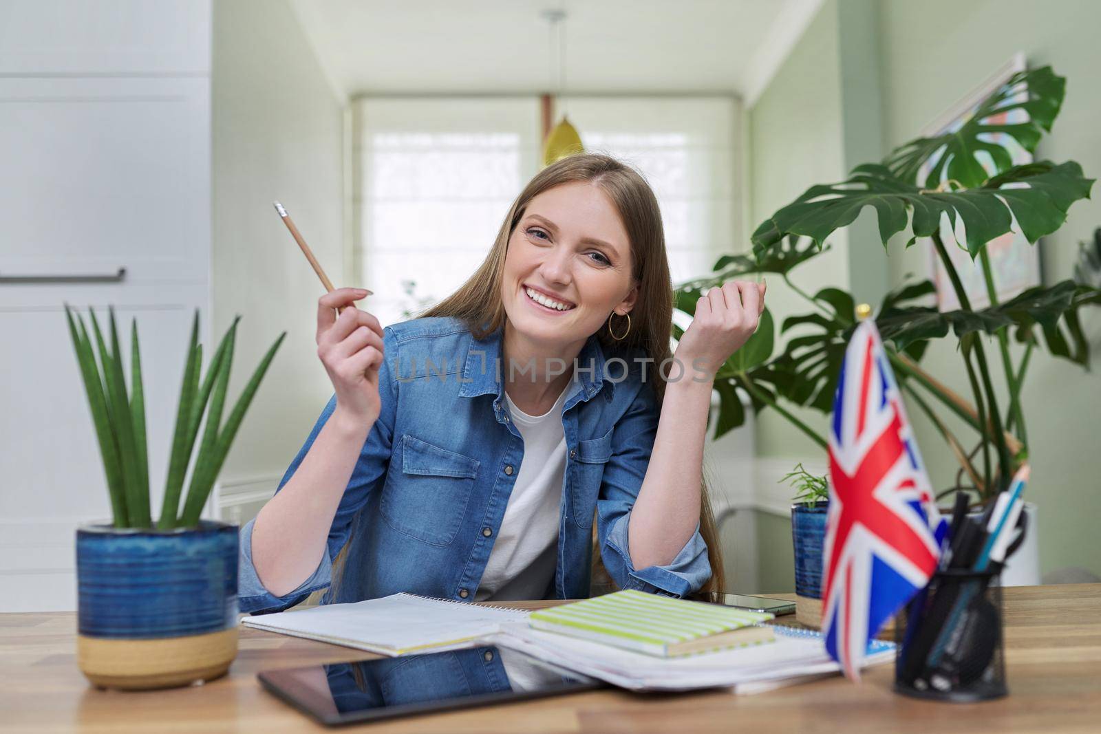 UK learning English online. Female student remotely looking at webcam taking private lessons sitting at home in kitchen, England flag background