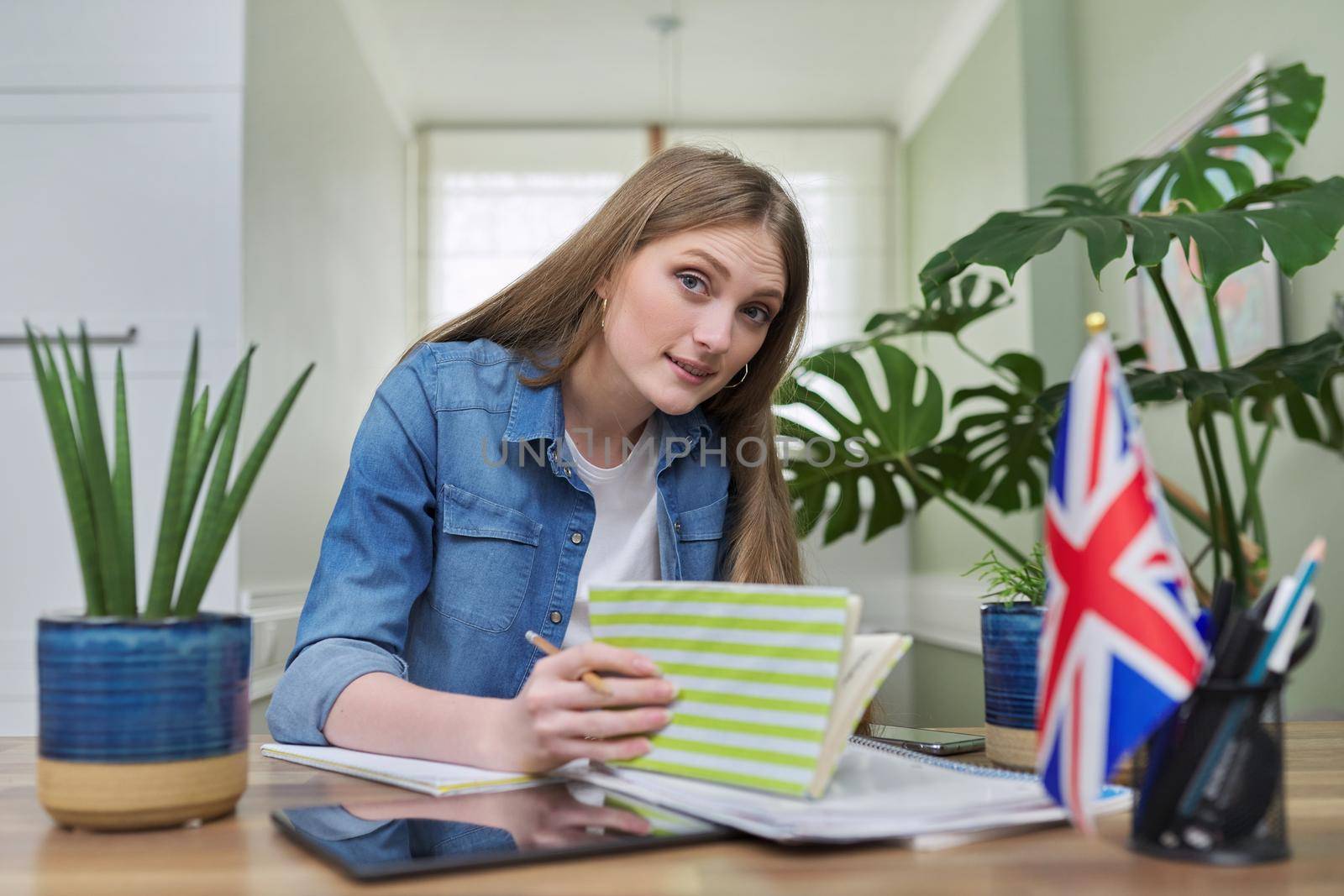 UK learning English online. Female student remotely looking at webcam taking private lessons sitting at home in kitchen, England flag background