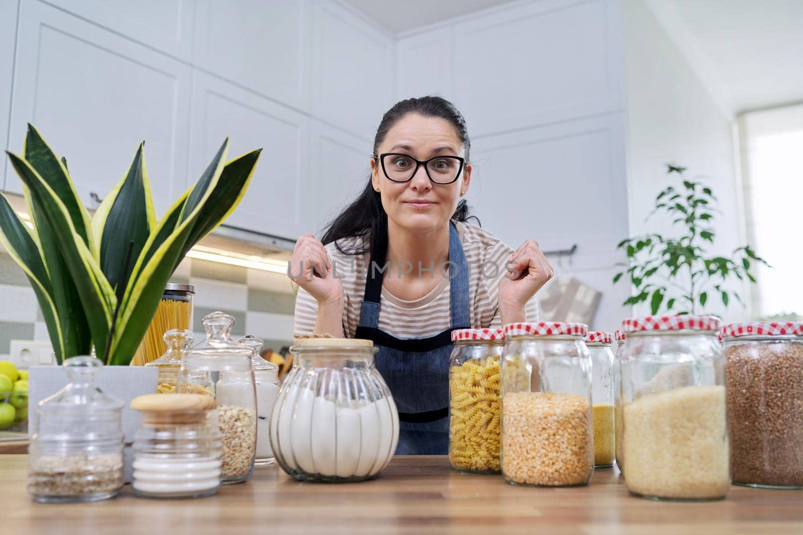 Storing food in kitchen, woman housewife in an apron with jars and containers of cereals, food, pasta, talking and looking at camera. Female blogging about organizing space in kitchen