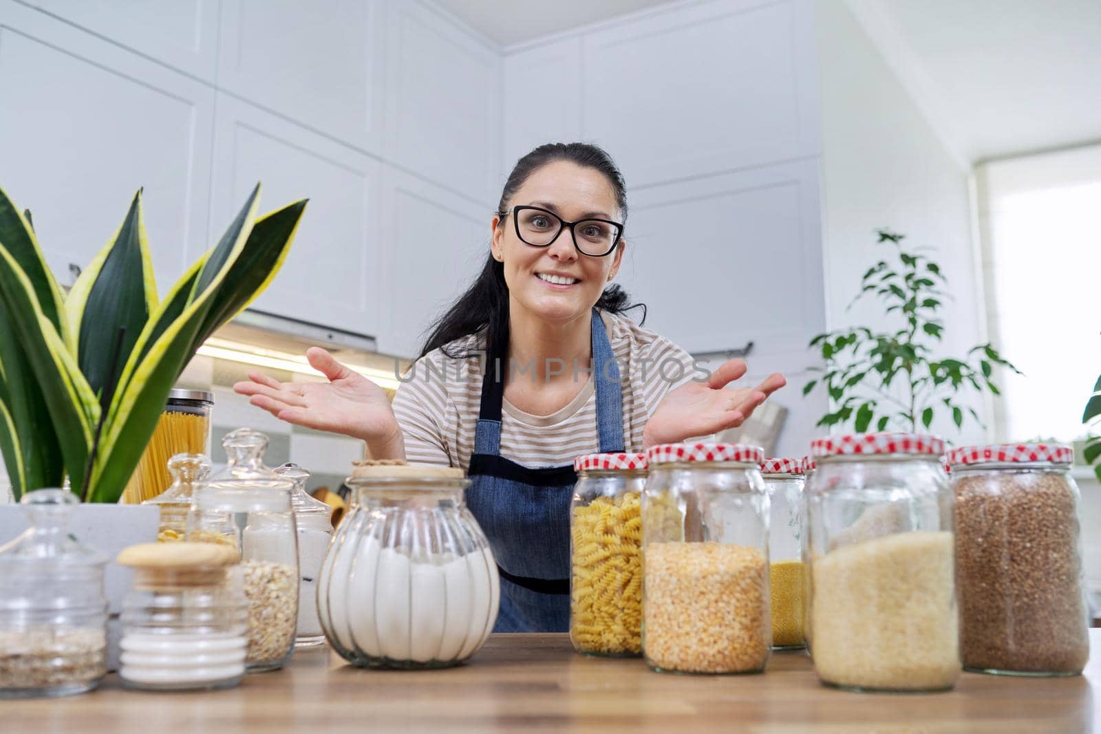 Storing food in kitchen, woman housewife in an apron with jars and containers of cereals, food, pasta, talking and looking at camera. Female blogging about organizing space in kitchen