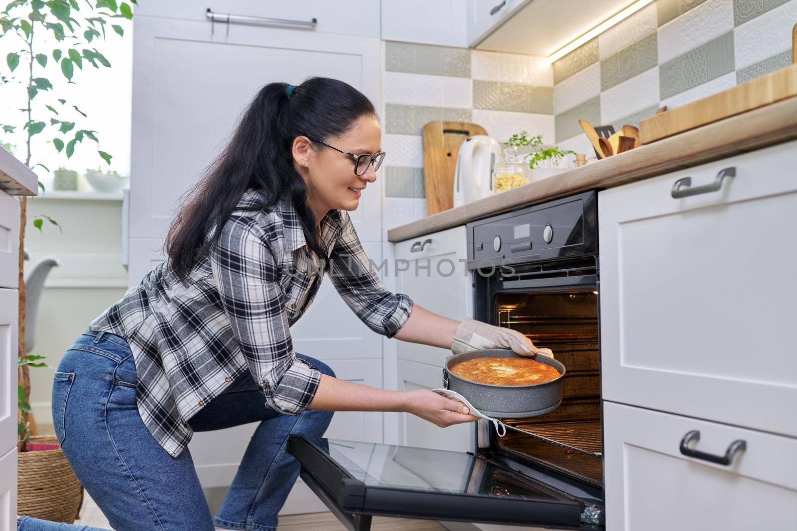 Woman at home in kitchen taking out hot apple pie from oven. Cooking at home, homemade baking, eating at home, lifestyle, hobbies and leisure