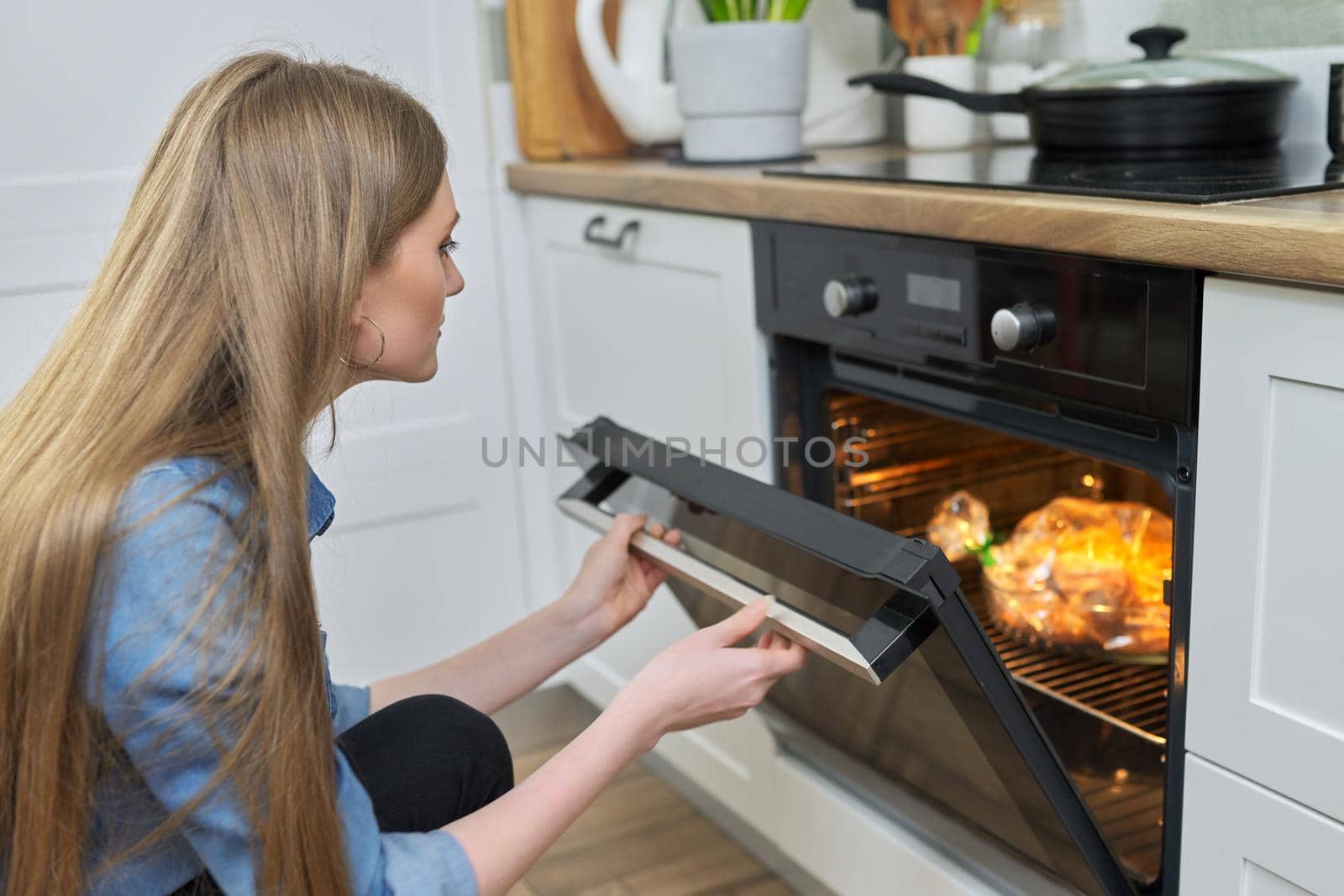 Cooking at home, roasting meat in oven, young woman putting marinated chicken in baking bag in oven, kitchen interior background