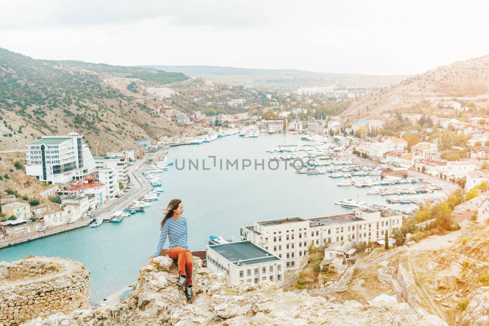 Beautiful young woman sitting on background of resort town around sea bay.