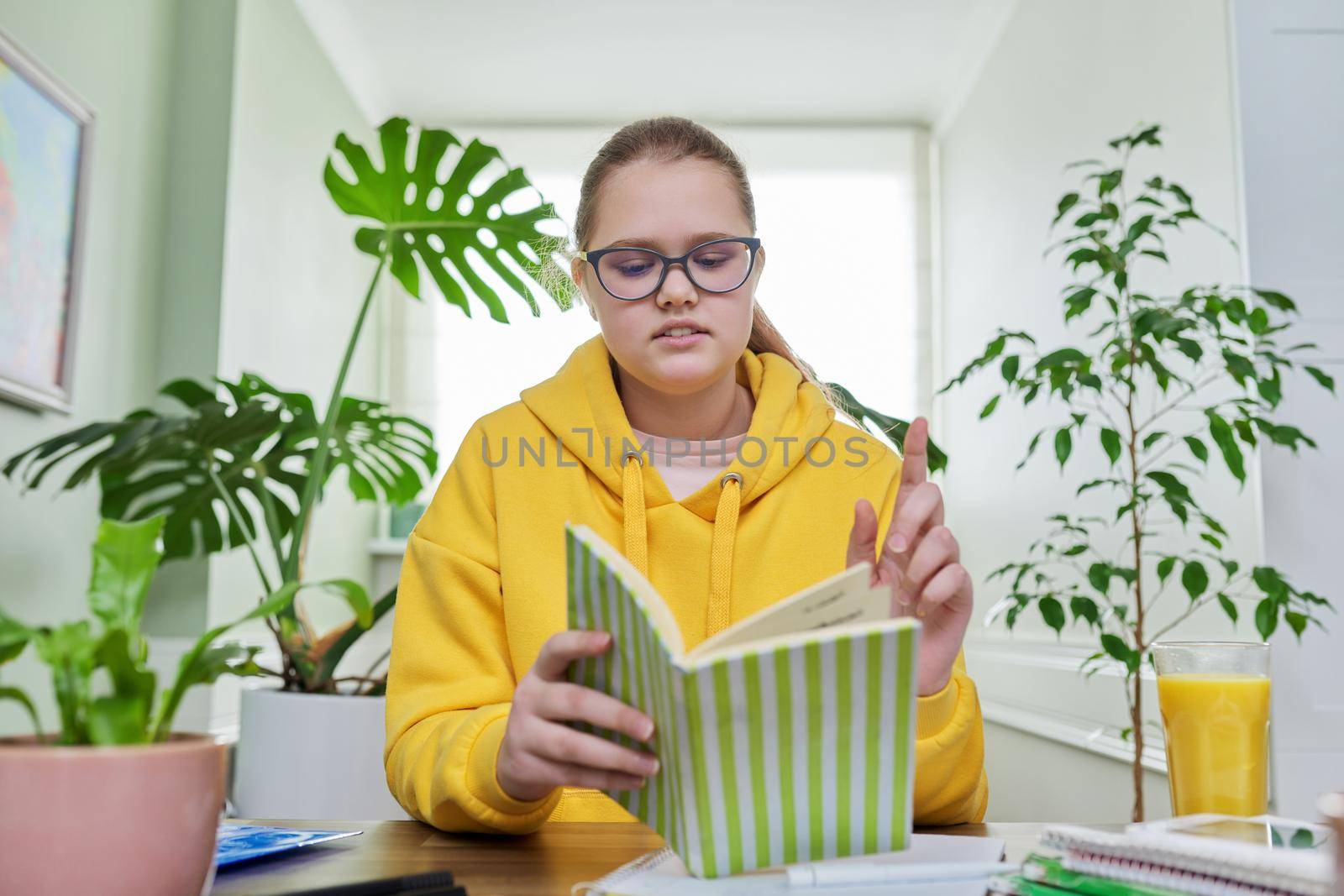 Pre-adolescent girl studying at home, talking schoolgirl looking at webcam, sitting at the table with books, textbooks, notebooks