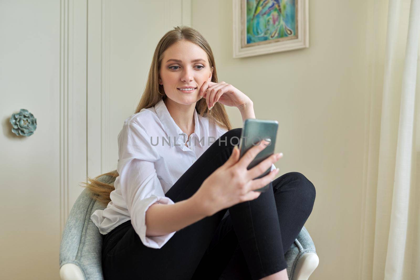 Young beautiful blonde sitting at home in chair, smiling talking using video communication on smartphone. Leisure, woman relaxing looking at phone screen