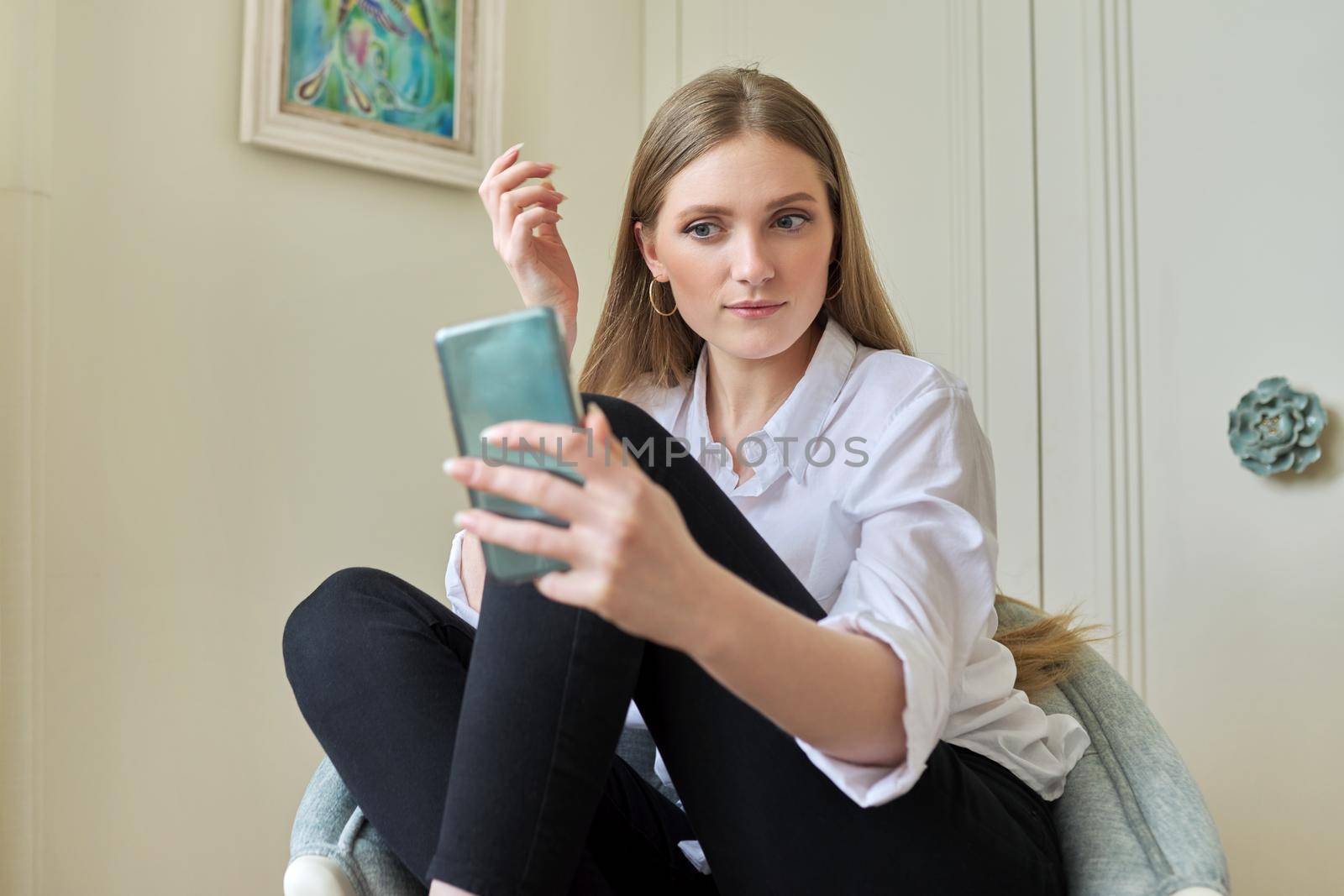 Young beautiful blonde sitting at home in chair, smiling talking using video communication on smartphone. Leisure, woman relaxing looking at phone screen