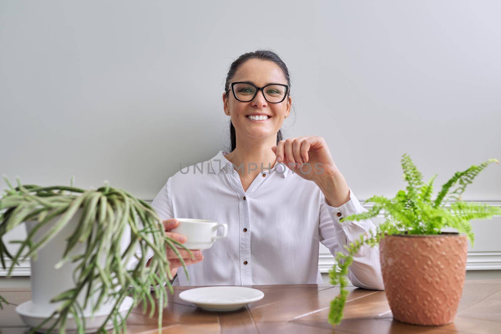 Talking positive business woman looking at camera. Middle-aged female wearing white shirt glasses sitting at table with cup of coffee, gesturing making video consultation, recording video vlog