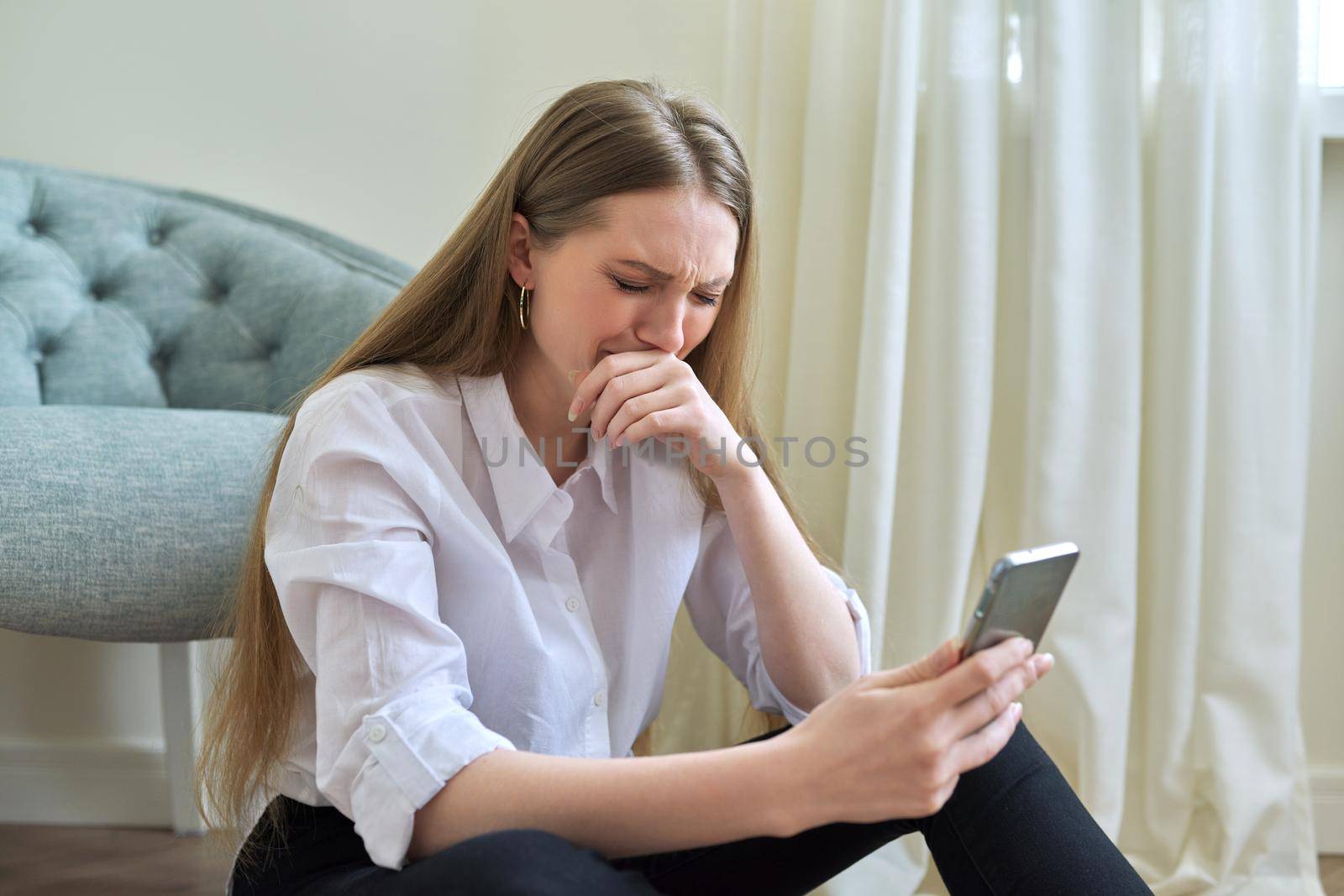 Sad crying young woman with smartphone sitting on the floor at home. Depression, bad news, problem, pandemic, illness concept