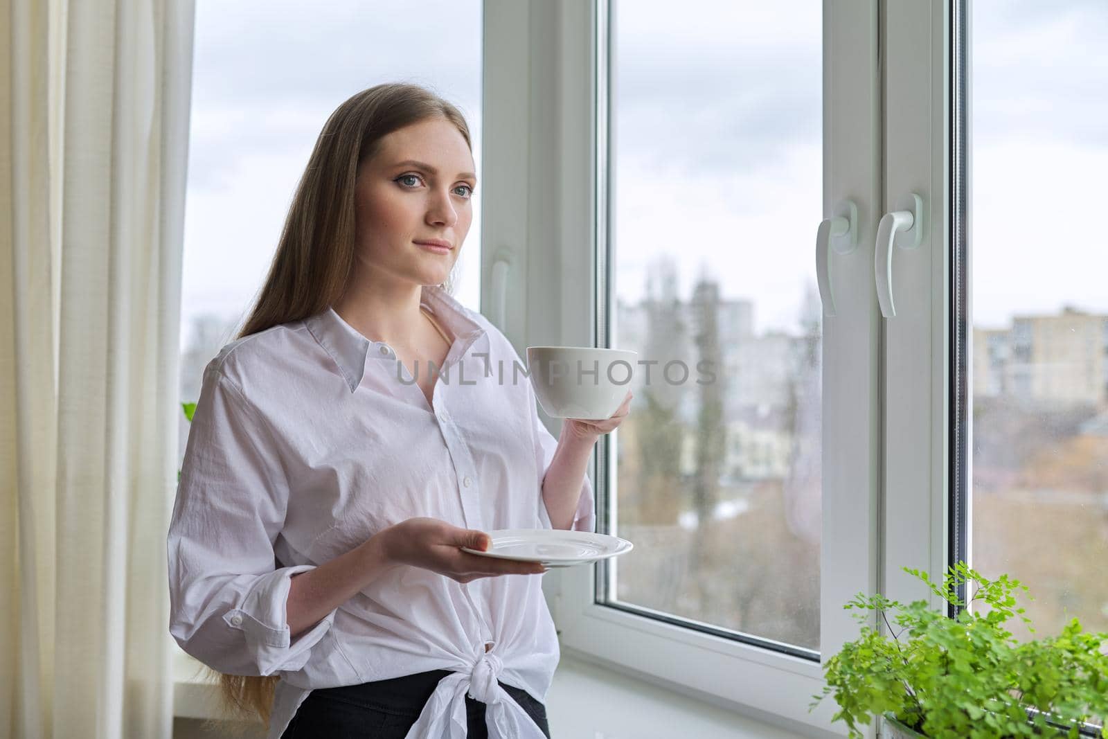 Beautiful smiling young woman with natural blond long hair holding cup coffee near panopam window. Attractive female in white shirt, winter spring season in window, copy space