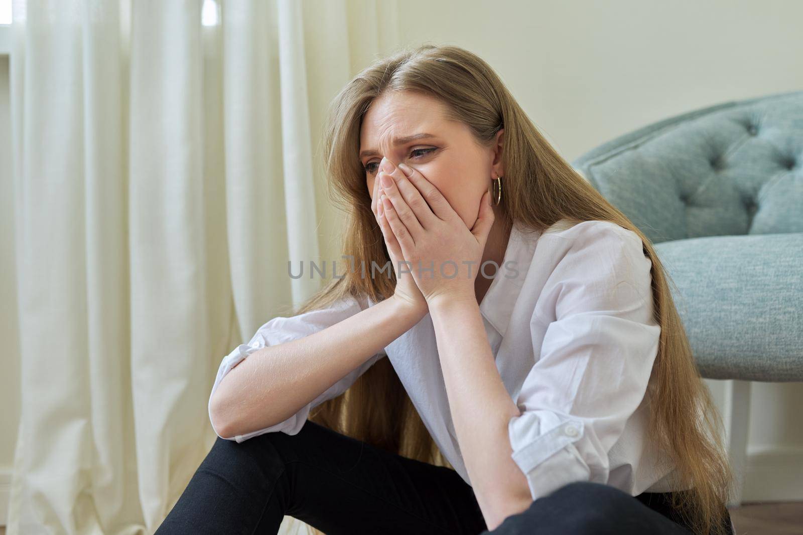 Upset crying young woman sitting at home on the floor. Negative emotions, stress, mental health, frustration, pain, people concept