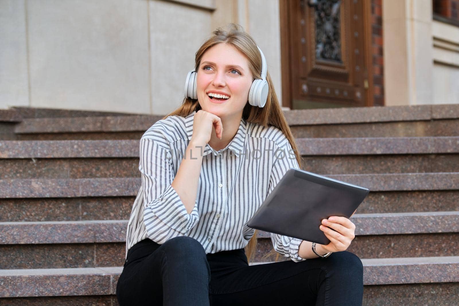 Young beautiful woman in headphones with digital tablet resting sitting on steps in city, listening music watching video. Happiness, relaxation, lifestyle, youth and technology