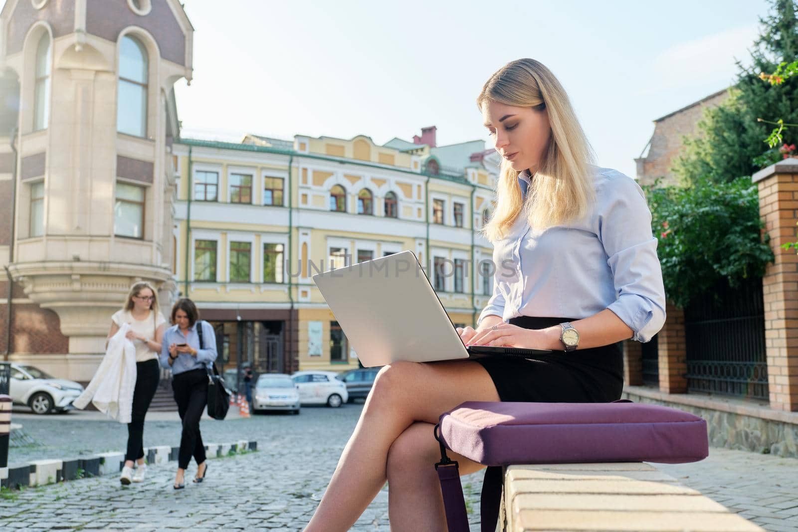 Beautiful young woman sitting outdoors using laptop, city street background. Female in business clothes, study, business, lifestyle concept