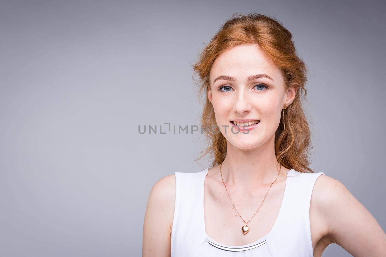 Closeup portrait young, beautiful business woman, student with lred, curly hair and freckles on face on gray background in the studio. Dressed in white blouse with short sleeves about open shoulders by Tomashevska