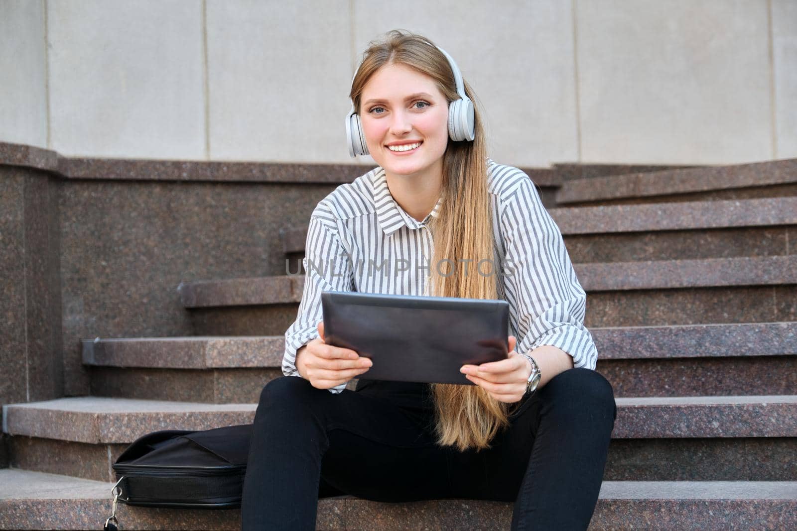 Young beautiful woman in headphones with digital tablet resting sitting on steps in city, listening music watching video. Happiness, relaxation, lifestyle, youth and technology