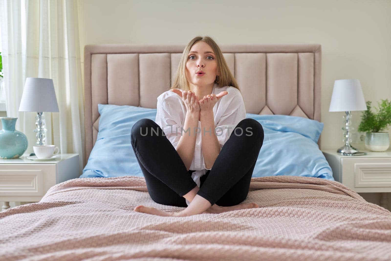 Lovely positive young woman showing air kiss gesture with hands and lips, beautiful blonde with long hair sitting in bed in pastel pink color