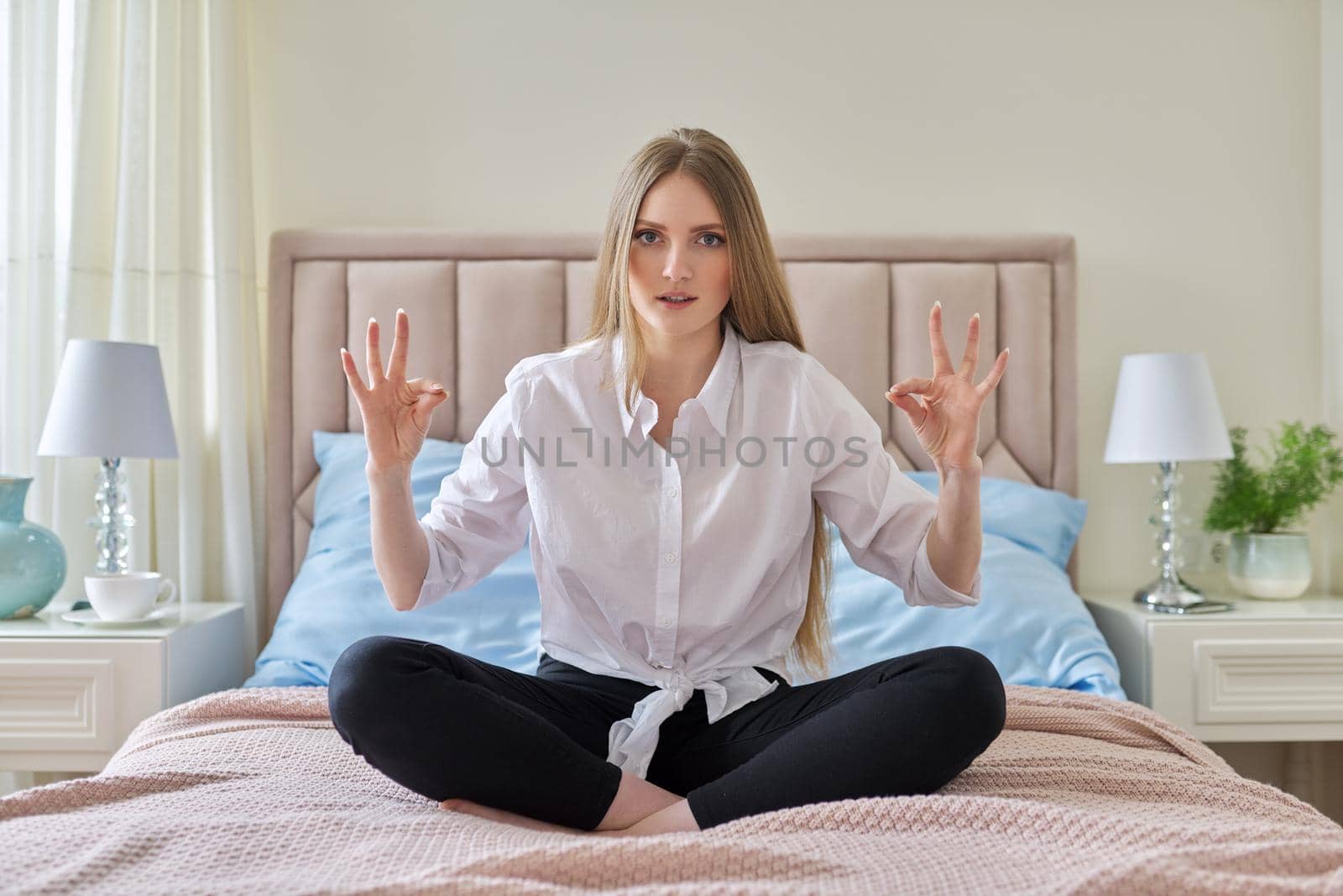 Yoga meditation at home, young beautiful blonde woman sitting in lotus position in bed, resting and meditating alone looking at the camera