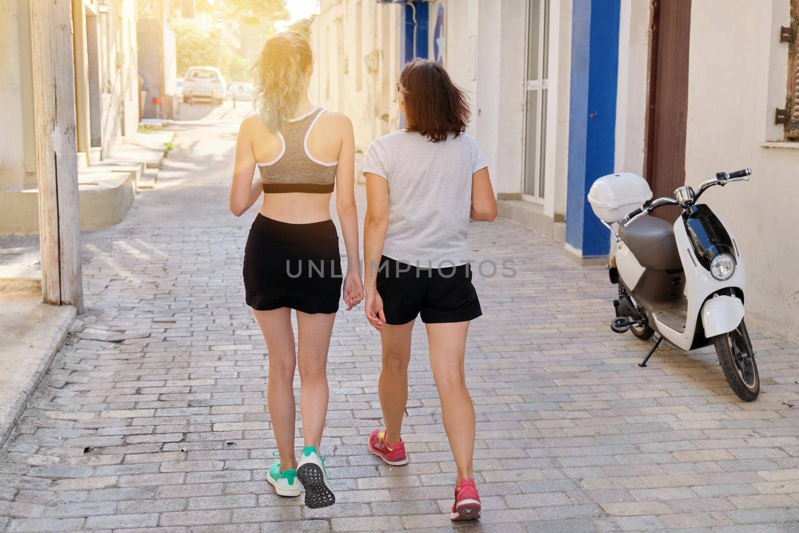 Mother and teenage daughter together in sportswear walking along the city street after fitness, jogging, back view