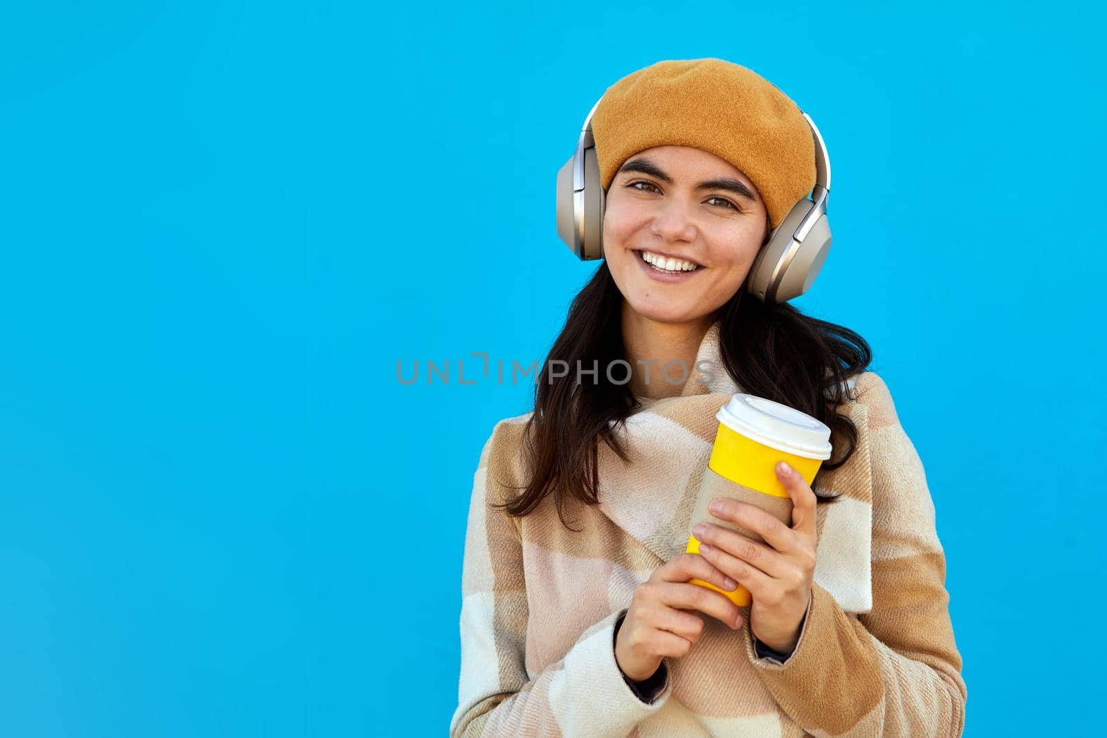 Cheerful stylish female in wireless headphones listening to music while standing with paper cup of drink to go on bright blue background
