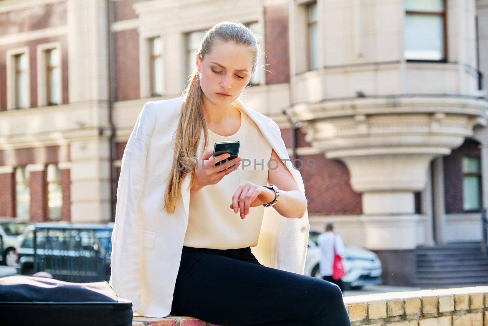 Young business woman in city looking at her wristwatch. Confident serious female with smartphone in her hands, sunset in the city, urban style background