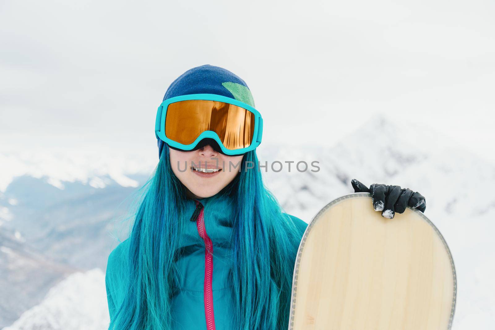 Smiling young woman with blue hair in sunglasses standing with snowboard in winter.