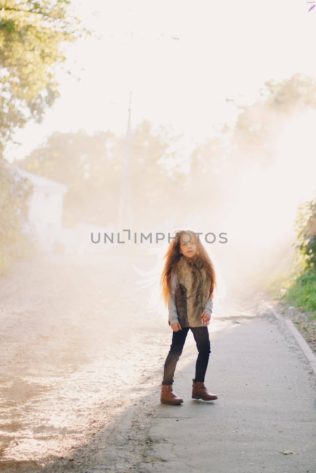 Stylish baby girl 4-5 year old wearing boots, fur coat standing in park. Looking at camera. Autumn fall season