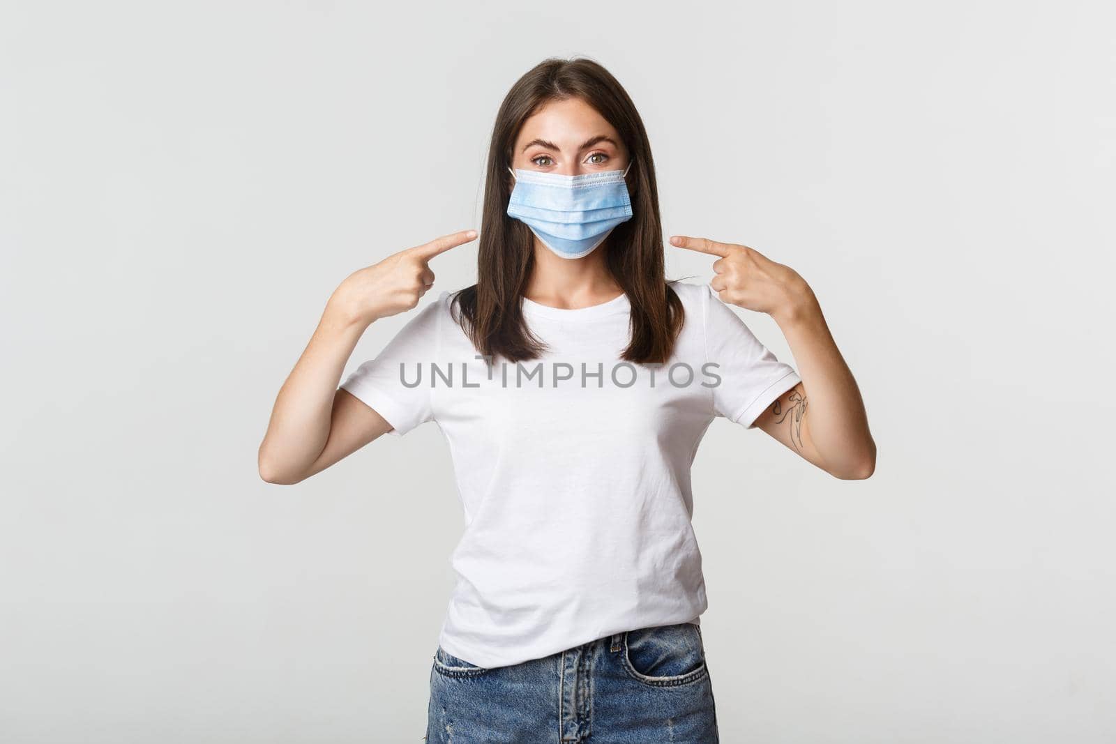 Covid-19, health and social distancing concept. Attractive brunette girl in medical mask pointing finger at face, white background.