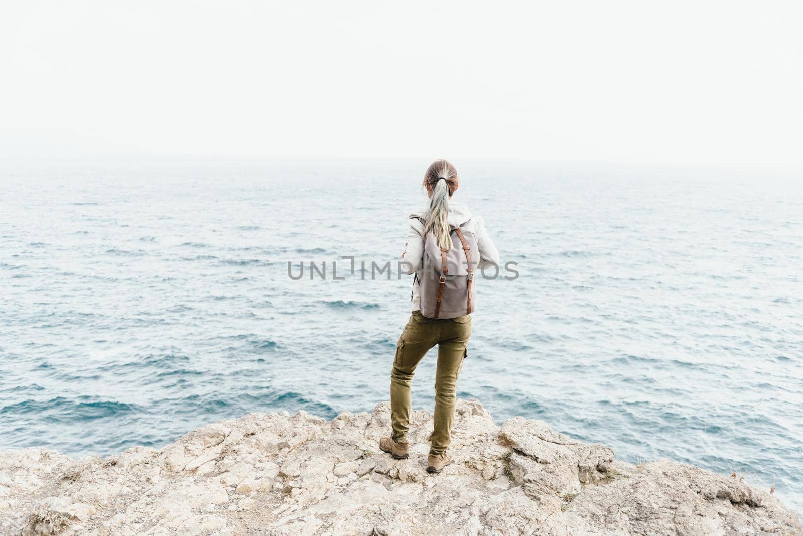 Traveler girl with backpack standing on rocky coastline and looking at sea.