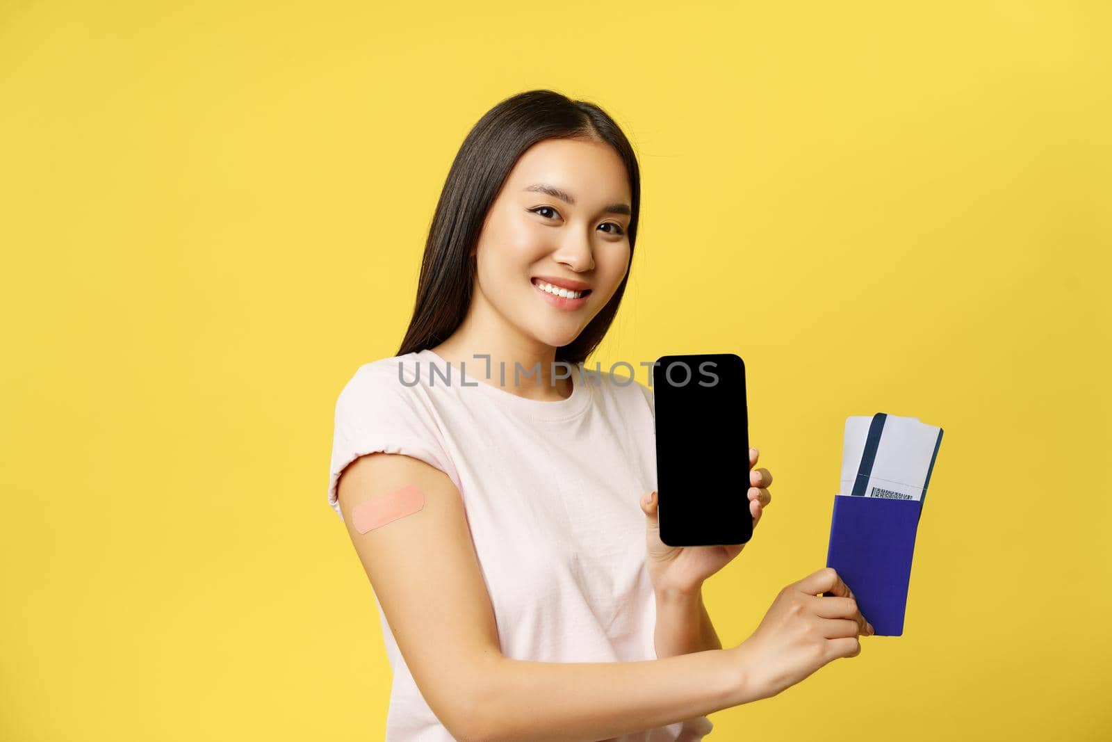 Smiling asian girl vaccinated, showing passport tickets and smartphone screen, app interface, standing over yellow background.