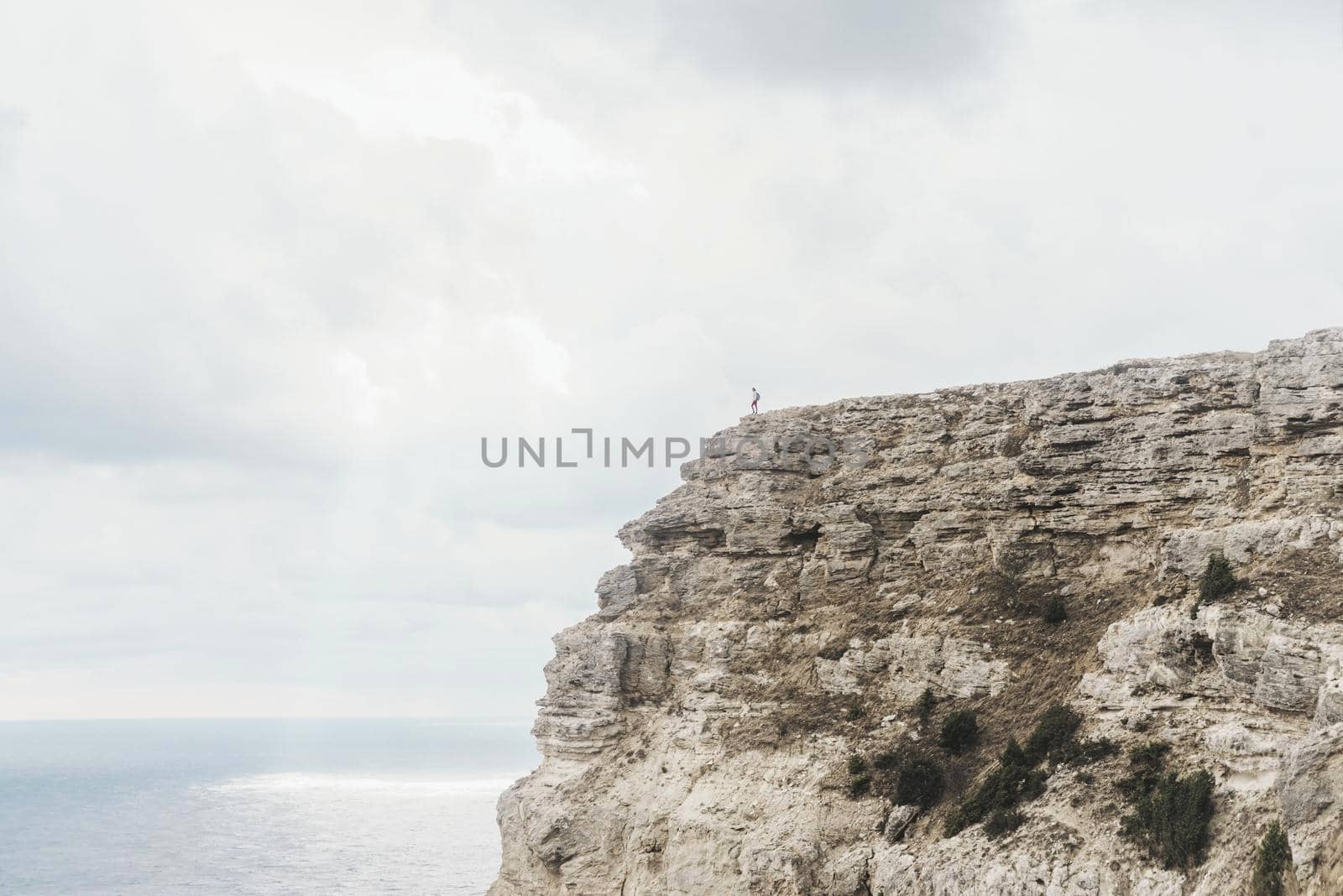 Small figure of female traveler standing on edge of cliff over sea on background of cloudy sky. Concept of minimal person in massive landscape.