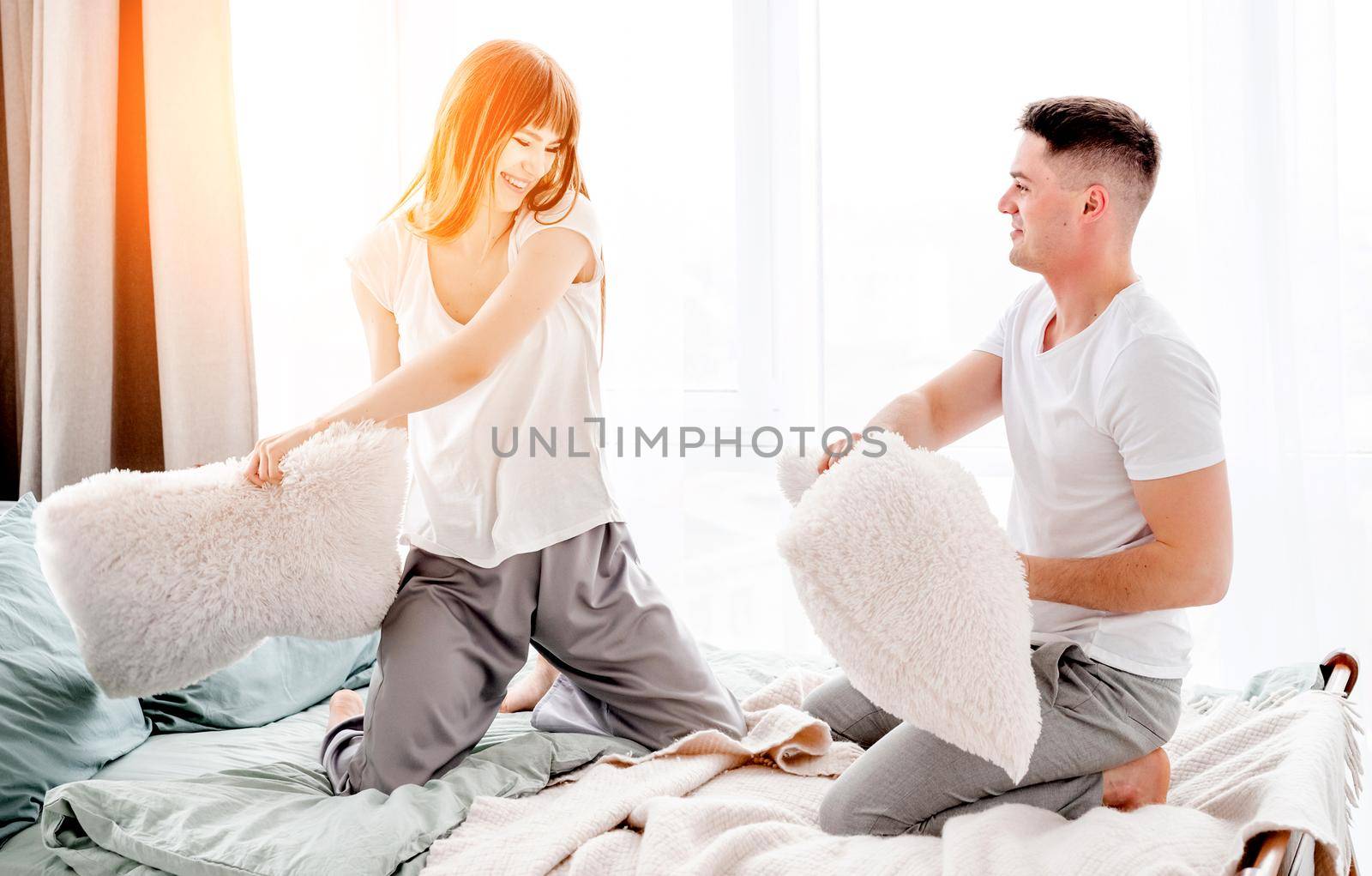 Beautiful young couple fighting with pillows in the bed and smiling. Girl and guy have fun in the bedroom with sunlight. Lovely family moments and enjoying of life